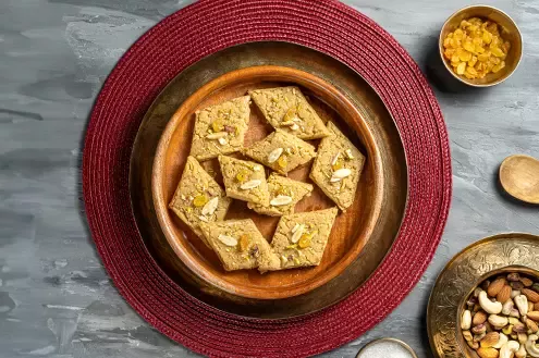 A wooden plate with pieces of a traditional sweet, garnished with nuts and dried fruits, on a red circular mat.