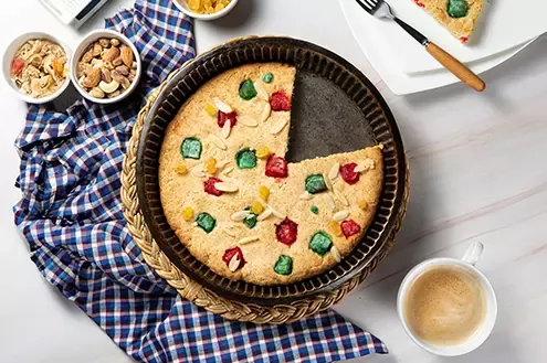 A colorful cake with dried fruits and nuts on top, partially cut with a coffee cup and bowls of nuts beside it