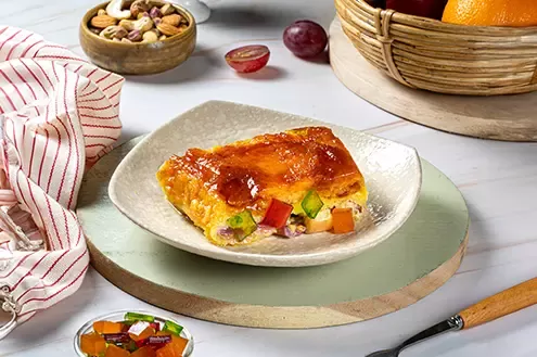 A square-shaped dish of baked dessert with mixed fruits, served on a modern plate with some fruit and nuts on the side.
